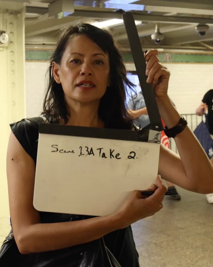 Martha Millan holding a slate marked Scene 13A Take 2 on a New York City subway platform.